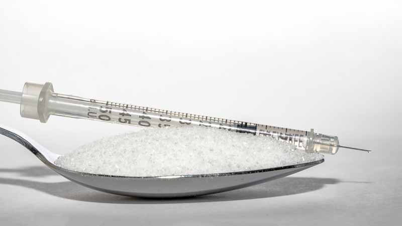 How to Stop Sugar Addiction - detoxandcure.com - image of a silver tablespoon on a white bench holding a full scoop of sugar with a hypodermic syringe resting on top of the sugar with the top of the syringe pointing out over the front of the sugar piled up on the spoon. The spoon and the syringe are casting a shadow on the bench and are in clear focus against a blurred white backdrop