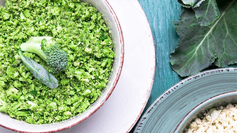 Keto Friendly Broccoli Fried Rice Recipe - white bowl with broccoli rice sitting on a turquoise timber table with a floret of broccoli in the middle of the bowl as a garnish