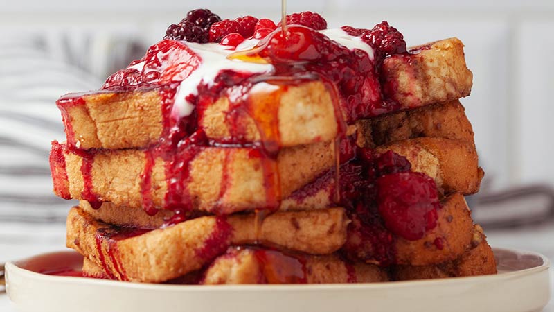 Delicious and Nutritious Sea Moss Vegan French Toast Recipe - Vegan French Toast stacked high on a round white plate topped with cherries and raspberries, Vegan cream, drizzled with Maple Syrup