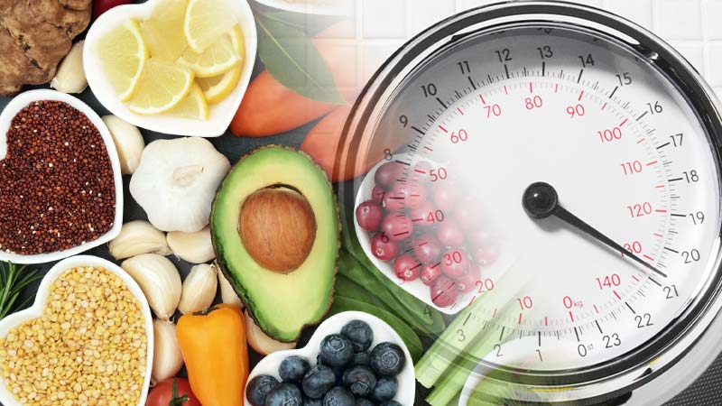 12 Tips for Losing Weight Fast at Home. Scales and a spread of whole foods on a stone bench top including avocado, cranberries, lemon, capsicum, carrot, garlic, bay leaves, ginger, lentils and tomato