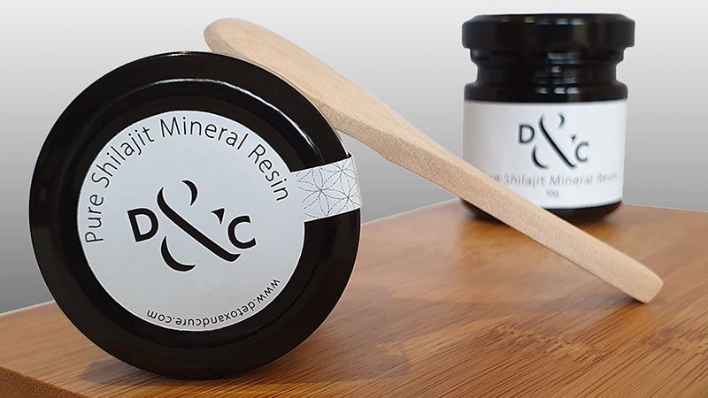 SHILAJIT BENEFITS; WHAT ARE THEY - image of a jar of 50g shilajit mineral resin laying face down on a timber board with a light coloured timber teaspoon leaning against it. In the background is a blurred jar of Shilajit Resin standing upright for visual effect