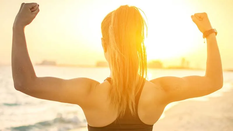 back facing shot of a young blonde woman with long hair standing in the sunset on a beach holding a strong pose with her fists up flexing her biceps. She is silhouetted against a city backdrop as the waves roll in to the front of her